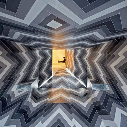 A person sits in a doorway at the end of a long, zigzag-patterned corridor. The tiling on the walls, floor, and ceiling showcases a geometric design with shades of gray. The doorway emits a warm, yellow light that contrasts with the cool tones of the corridor.