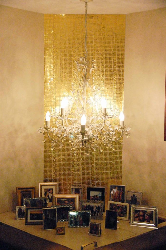 A corner hung chandelier backed with a gold mosaic wall reflecting the light of the chandelier beautifully